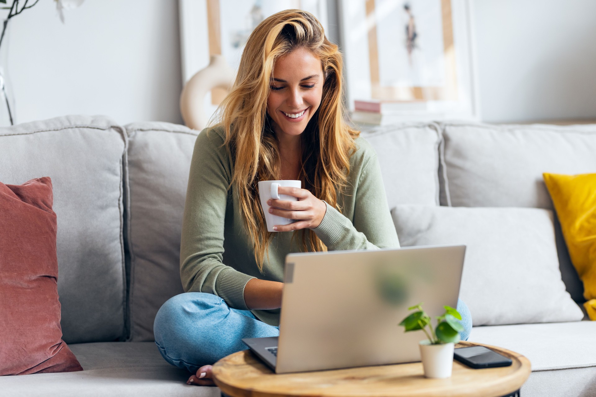 Beautiful young woman working with her laptop while drinking a cup of coffee sitting on a couch at home